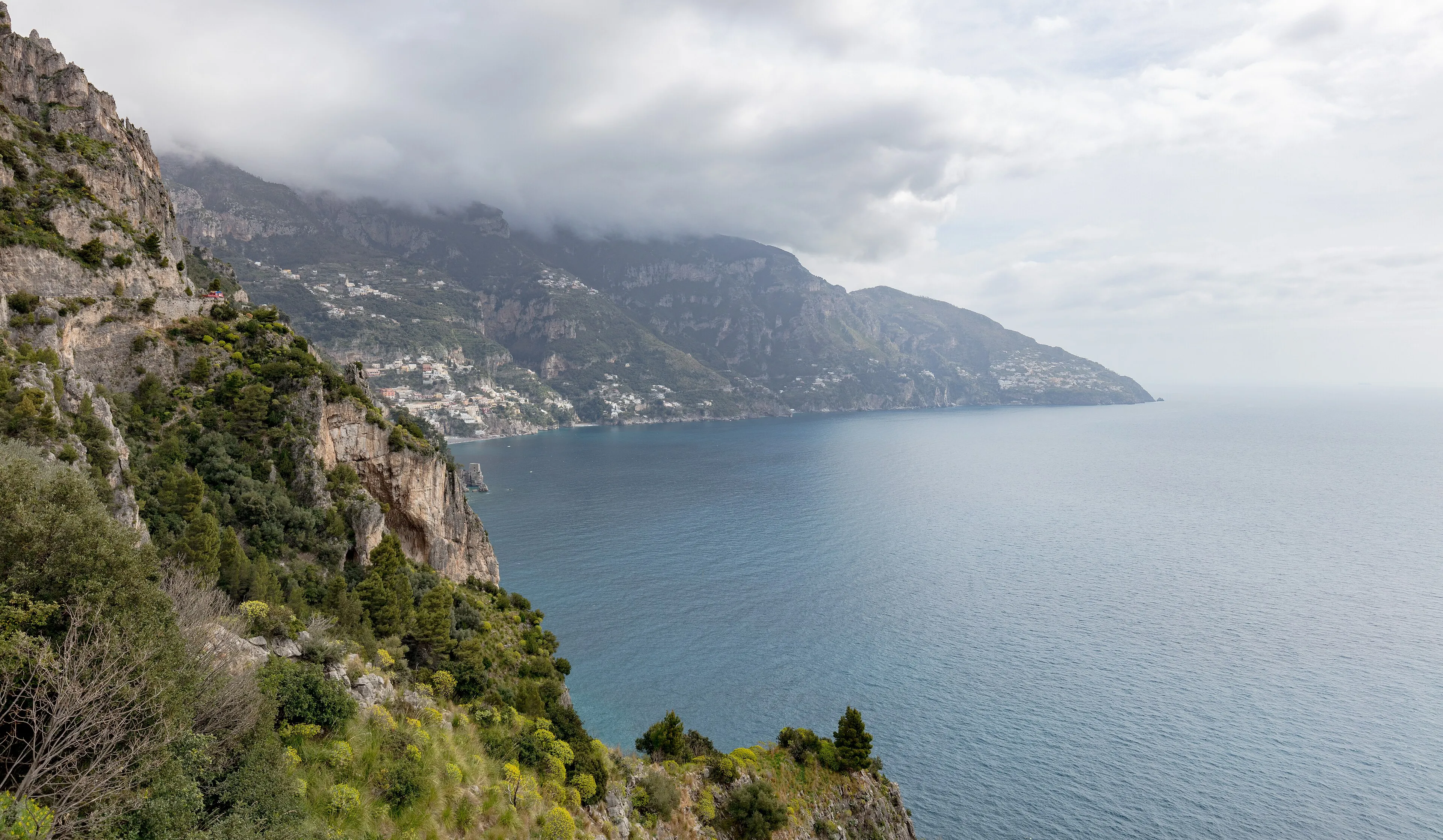 Rugged green cliffs above the sea on the Amalfi Coast, with Positano small in the distance under low cloud