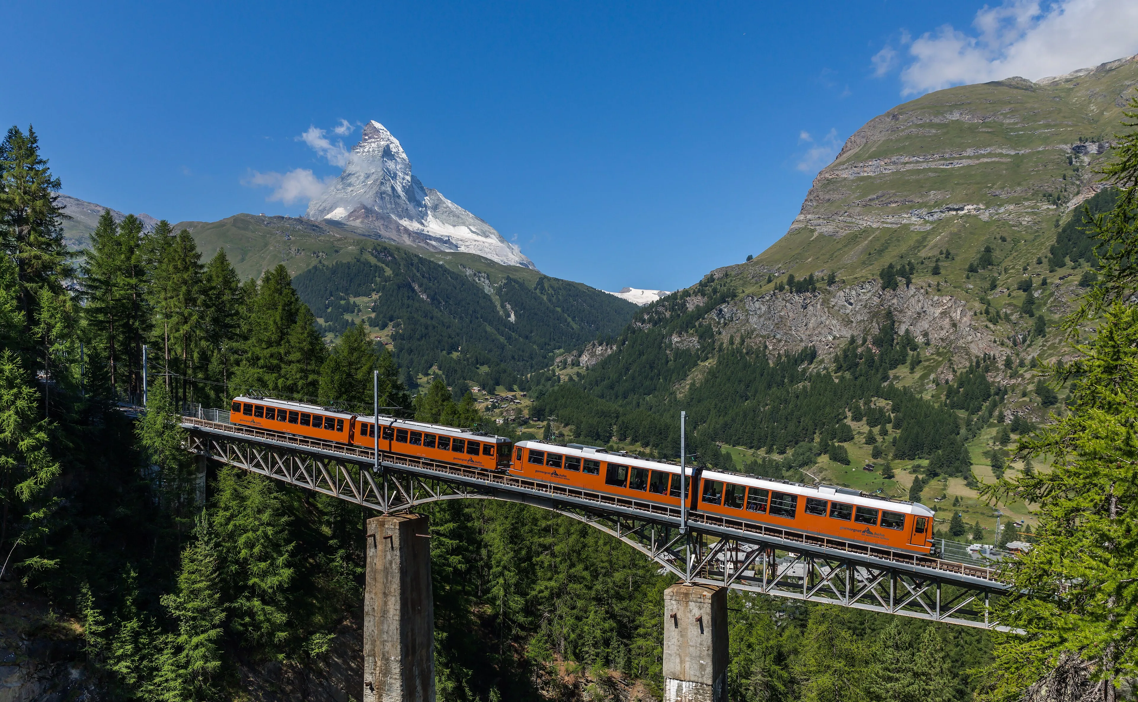 An orange Gornergrat railway train crossing a high bridge through the forest above Zermatt, with the Matterhorn beyond