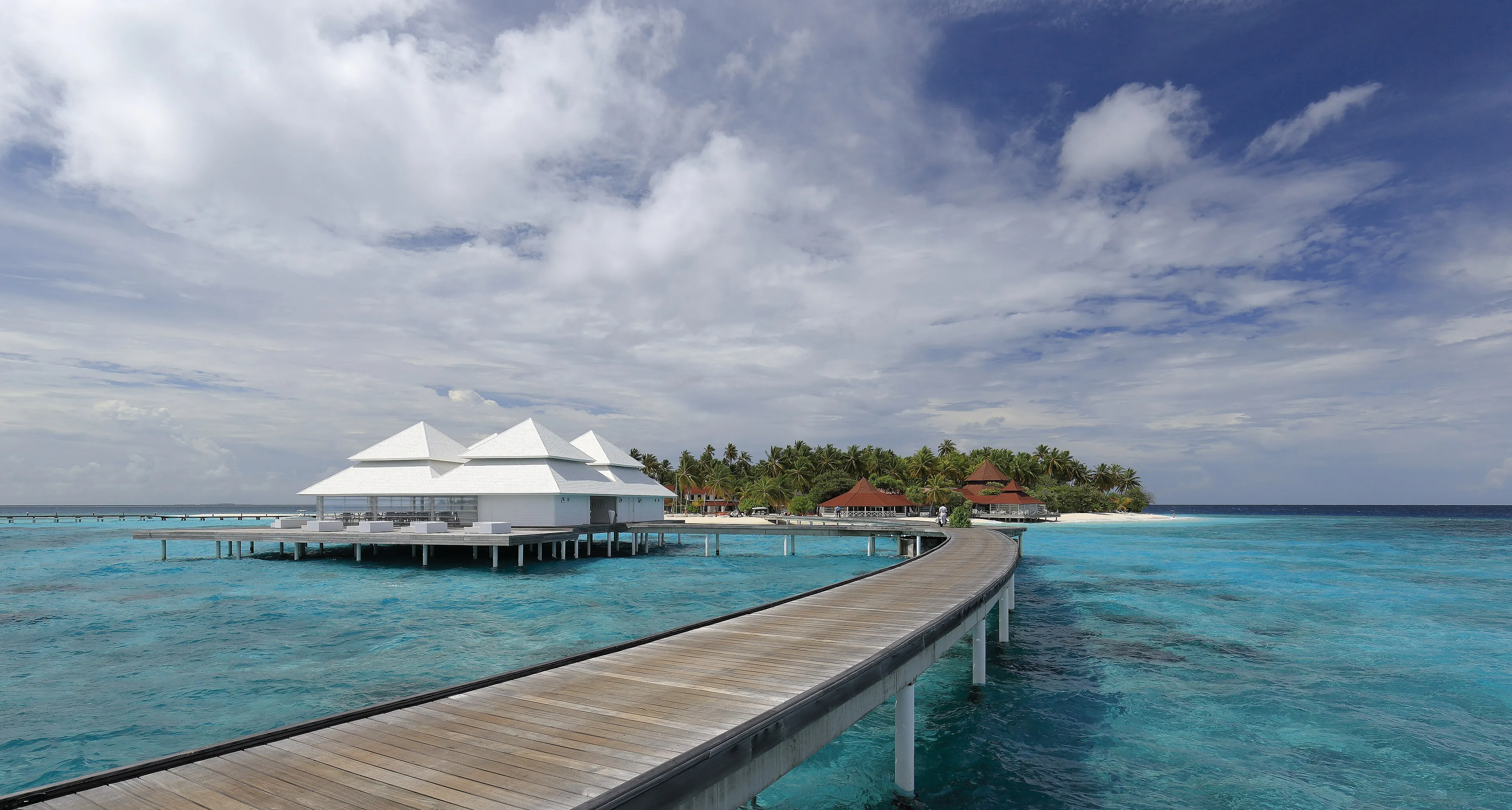 A curving wooden jetty over turquoise water leading to a white overwater pavilion and a palm-covered island