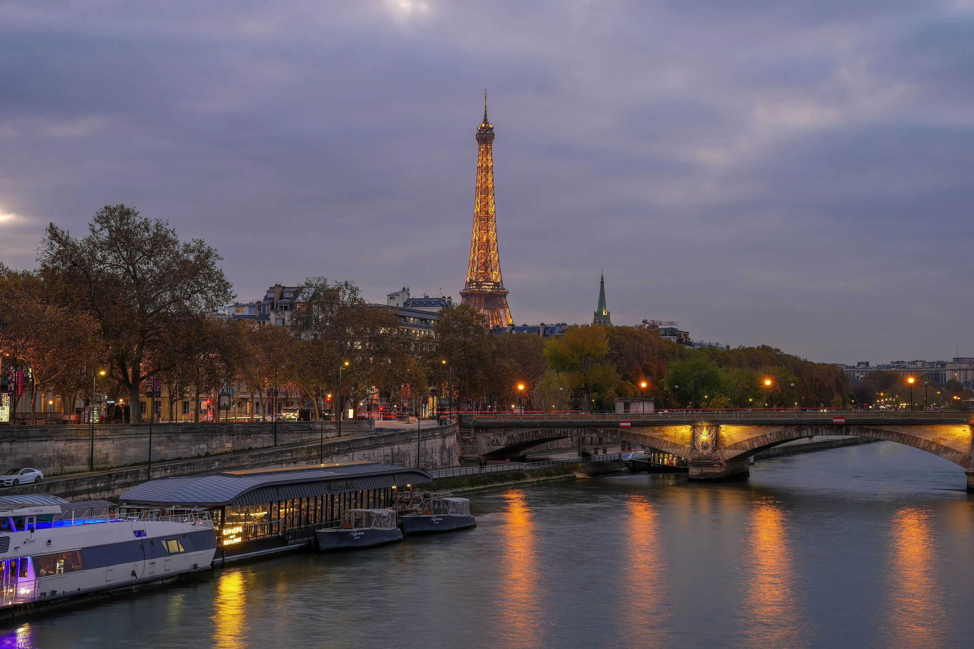 The Eiffel Tower lit up at dusk above the Seine, with river boats moored along the bank