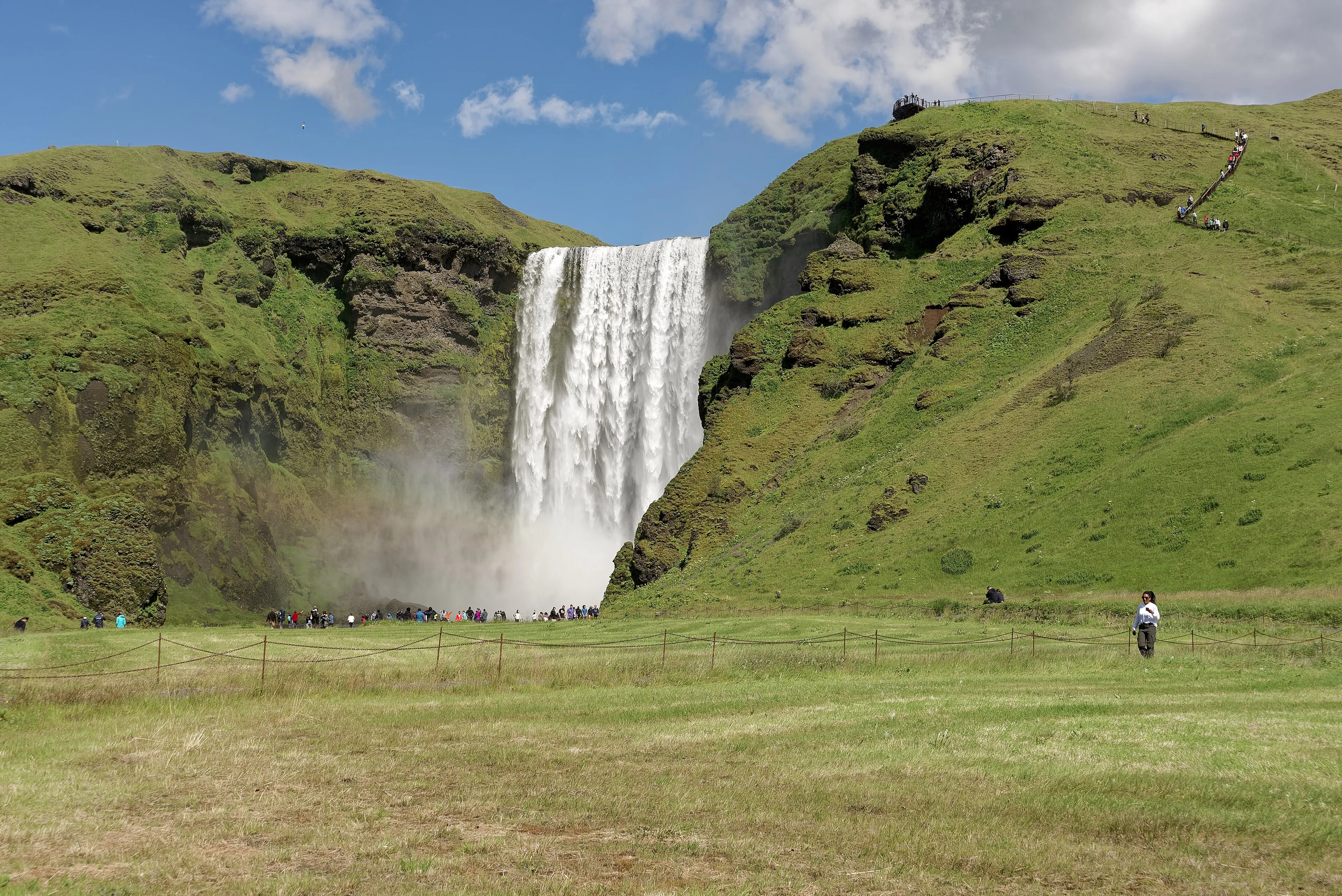 Skógafoss waterfall plunging from a green cliff on Iceland's south coast, with visitors small at its base
