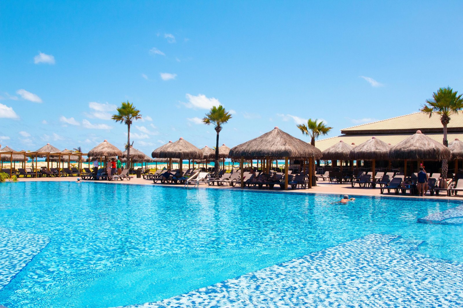 A beautiful tropical resort featuring a pool, thatched huts, and palm trees under a clear blue sky.